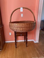 Front view of very large round wicker basket with a thick twisted handle, placed on a wooden stand against a salmon wall.