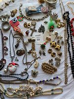 Top down angled photo of an assorted collection of costume jewelry including necklaces, earrings, pendants, and brooches laid out on white sheet.