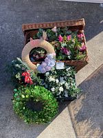 Photo showing the large woven wicker basket open with multiple faux floral wreaths and garland inside. The wreaths include a green leafy wreath, a burlap-wrapped wreath with white/lilac flowers and a butterfly, and a floral garland with pink flowers.