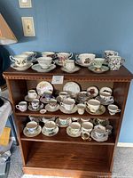 Wide view of multiple teacups and saucers arranged on three shelves of a wooden shelf unit against a blue wall, showing a variety of patterns and styles.