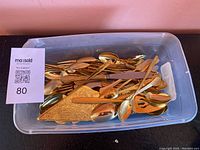 Top view of clear plastic container holding multiple assorted gold-tone flatware pieces including spoons, serving utensils, and knives.