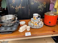 Overview of various kitchen items on a wooden table including baking pans, ceramic plates and mugs, small appliances.