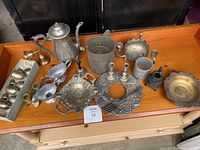 Overview of the mixed metal items on wooden table, showing teapot, tray, candle holders, small bowl, goblets in box, and other items.