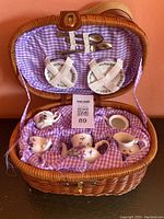 Porcelain doll-sized tea set arranged inside open wicker picnic basket lined with purple gingham fabric. Shows teapot, creamer, sugar bowl, two cups and saucers, plates, and metal toy cutlery held in loops on basket lid.