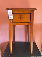 Front view showing small pine side table with single drawer and knob on a pink background.