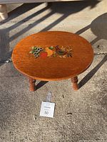 Small wooden oval foot stool with hand-painted colorful fruit motif on top, four turned legs visible, photographed outdoors on concrete surface with sunlight and shadows.