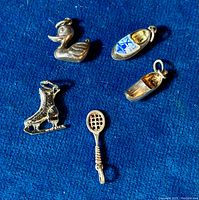 Set of five sterling silver charms on a blue background: a duck, Delft-pattern shoe, ice skate, tennis racket, and wooden clog.