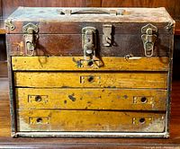 Front view of antique wooden tool chest with three yellow drawers and metal latches, showing wear and paint chipping.