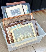 View of framed paintings and prints with visible frame styles and painting subjects in a plastic storage bin