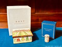 Photo showing the larger square trinket box with countryside scene and the smaller cylindrical enamel box next to original Halcyon Days boxes on blue fabric.