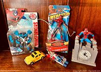 Transformers Robots in Disguise Blizzard Strike Optimus Prime and Spider-Man Web-Riders toy figures, with yellow Volkswagen Beetle car, red/silver airplane, and Spider-Man clock tower figure laid out