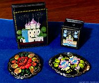 Photo showing four vintage hand-painted black lacquer items on a blue background: two rectangular trinket boxes decorated with Russian architectural motifs and two oval floral brooches.