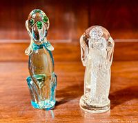 Pair of hand blown Murano glass dog figurines: taller blue-tinted dog with green eyes and nose, sitting upright; smaller clear glass dog with textured internal pattern, both on wooden surface.