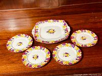 Five plates arranged on wooden surface: four round plates and one square plate with handles, all with colorful floral pattern on white porcelain.
