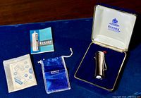All contents displayed on blue surface: presentation case open with lighter, velvet pouch, instruction booklet and sheet