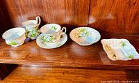 Full view of five bone china pieces on wooden shelf showing creamer, sugar bowl, teacup with saucer, bowl, and divided candy dish, all with green trillium floral pattern.