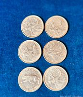 Six Canadian silver quarters laid out on blue fabric, reverse side visible featuring caribou figure.