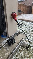 Side view of a red and black Homelite 2-stroke trimmer leaning against a wall outside on stone steps with light snow on the ground.