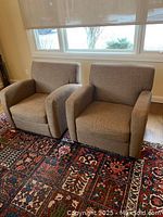 Pair of light beige fabric club chairs set on a hardwood floor near a window with a patterned carpet underneath.
