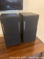 Pair of JBL HLS610 speakers with black grilles installed, placed on wooden surface.