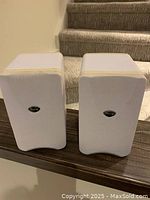 Two white Mirage speakers with cloth grille covers displayed side by side on a wood surface with stairway background.