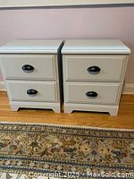 Front view of the pair of small white nightstand bedside tables with black drawer pulls, showing both tables side by side on a hardwood floor with a patterned carpet in front.