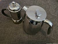 Photo showing stainless steel Bodum French press coffee maker next to a stainless steel Bodum tea pot, both placed on a dark fabric surface.