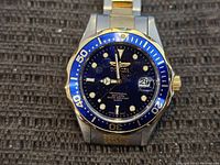 Close-up front view of men's Invicta wristwatch showing blue dial with luminous markers, date window, and two-tone silver and gold bezel and band.