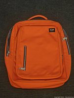 Full view of the orange Jack Spade backpack showing front zippered compartments and top handle.
