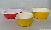 Three vintage Pyrex bowls in red and yellow with white interiors shown side by side on a plain background.