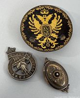 Photo showing the three items together: the round German Empire coat of arms mini plate, the Scottish clan badge, and the Victorian oval brooch.