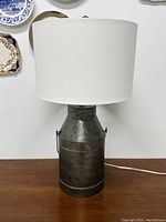 Front view of the metal milk can style table lamp with white drum lampshade on wooden surface.