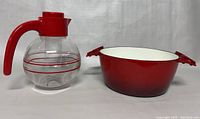 Photo showing side view of clear glass pitcher with red handle and red striped design, next to red cast iron pot with white enamel interior and two handles.