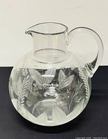 Photo of clear round glass pitcher with etched floral and leaf design and curved handle on black surface to show overall appearance and condition.