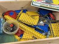 Overall view showing a box filled with various Meccano parts such as perforated metal plates, red and blue wheels, and a small container with screws and bolts.
