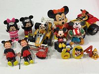 Various Disney character toys including multiple Mickey and Minnie Mouse figures, toy vehicles, and small character figurines arranged on a white surface.