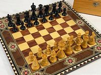 Chess board set up with all chess pieces in starting positions, showing intricate decorative inlay on border and full set of black and natural wood pieces.