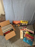 Full view of cardboard box packed with approximately 60 vinyl records plus several records positioned outside the box showing various album covers including The Association's 'Birthday' and Kinks single 'Kinks-Size'.