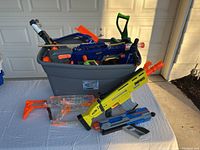 Various Nerf toy blasters including clear plastic blaster, Fortnite-themed blaster, and other assorted styles and colors in a large storage bin and on table.