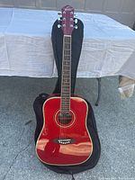 Red Oscar Schmidt by Washburn acoustic guitar with black gig bag underneath, pictured outdoors on concrete with a table in background.