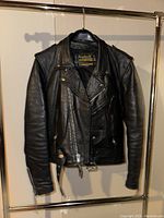 Front view of the black leather biker jacket hanging on a metal rack, showcasing zipped pockets, asymmetrical zipper closure, and belt at the waist.