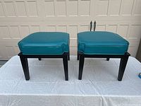 Two square black frame footstools with teal cushions on white table