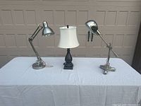 Front view of the set showing two adjustable metal desk lamps and one traditional table lamp with black base and white shade on a white cloth-covered table.