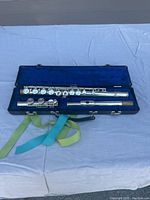 Flute disassembled into three parts lying in an open blue velvet-lined black case with carrying handle and green and blue strap.