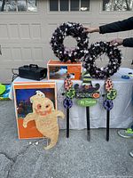 Photo of all items on table: fog generator, boxed ghost with lights, two metallic purple and silver wreaths, two wooden yard signs with Halloween themes