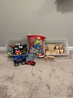 Wide shot of the lot showing three separate plastic bins: one holding Mega Bloks in a red container, one with various small toy cars and figures, and one with wooden blocks including magnetic ones; with a couple of toy vehicles in front of bins.