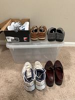 Full view of five pairs of men's shoes arranged on and in front of a plastic bin: white and blue Under Armour athletic shoes in box, tan boat shoes, grey and black slippers, white Nike Air Monarch sneakers, and burgundy loafers.
