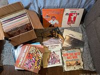 Cardboard box filled with approximately 75 vinyl records showing edge of album covers; some loose records arranged outside the box.