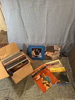 Photo of a cardboard box filled with approximately 50 vintage country vinyl records and several album covers displayed beside it including Eddy Arnold, Hank Williams, Charlie Rich, and a blue cover with a couple.