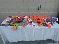 Multiple Nerf guns including rifles, pistols, and accessories laid out on a table; mostly orange and white.